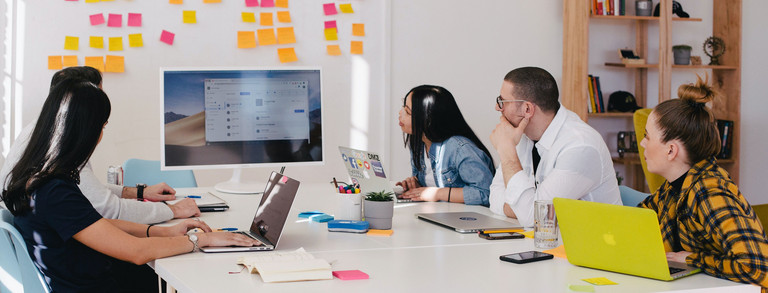 5 Personen sitzen um einen Tisch und schauen gemeinsam auf einen Bildschirm. Alle haben Laptops und Arbeitsmaterial vor sich und im Hintergrund steht ein Whiteboard mit verschiedenfarbigen Post-Its.