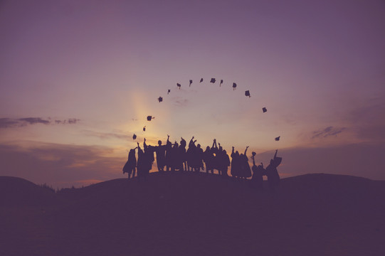 The sun rises behind a hill. A line of people stands on the hill, their silhouttes dark against the sunrise. People have thrown their university graduation hats in the air at the same time.