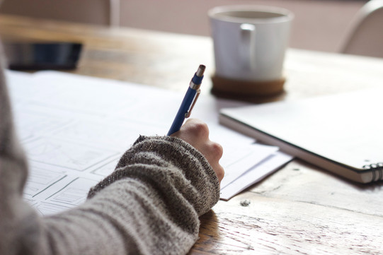 Writing A hand is writing with a ballpoint pen on the sheets of paper in front of it. On the table where the person is working, there is also a writing pad and a cup.