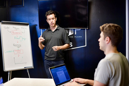 One man is standing at a flip chart with a process diagram explaining something to another man sitting at a laptop.