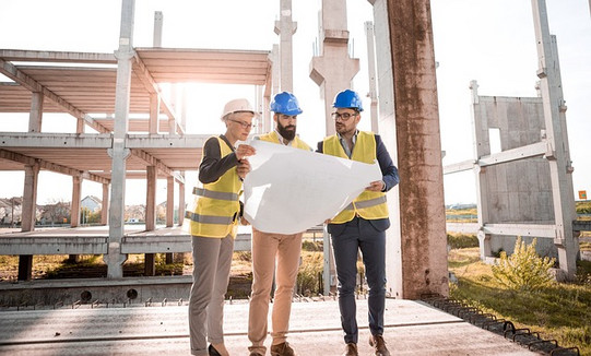 A woman and two men wearing safety helmets and safety vests are standing on a construction site studying a large plan.