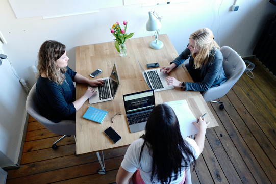 Drei junge Frauen sitzen an einem Tisch, vor ihnen jeweils ein eingeschalteter Laptop, Handys und Schreibutensilien. Die Frauen sind im Gespräch.