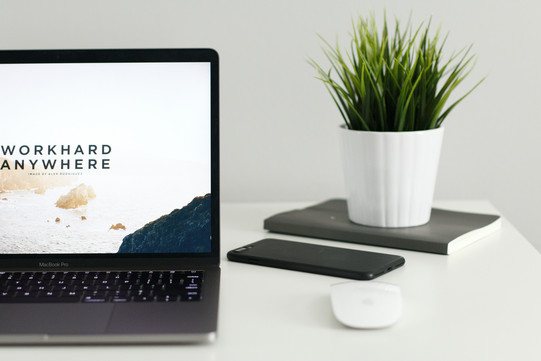 An open laptop with "Work hard anywhere" written on the screen. Next to it on the table are a PC mouse, a cell phone, and a notebook with a plant on it. 