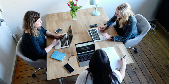 Three young women are sitting at a table, each with a switched-on laptop, cell phones and writing utensils in front of them. The women are in conversation.