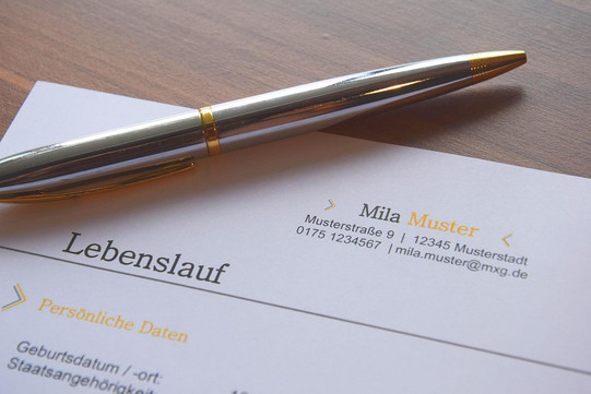 Upper third of a sample CV on paper. You can see the sample address, the German word for CV and the beginning of the personal data category. On top of it is a silver ballpoint pen.