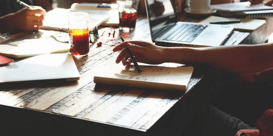 Several people are sitting around a table. On the table an open laptop, open notebooks and drinks.