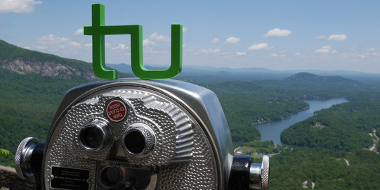 Blick an einem Aussichtsfernrohr vorbei auf eine schöne grüne Landschaft, durch die sich ein Fluss schlängelt. Auf dem Aussichtsfernrohr steht das TU-Logo.
