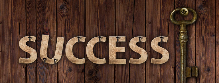 Auf einem Hintergrund auf Holzbrettern ist mit Nägeln das Wort Success in Großbuchstaben angebracht. Dahinter ist hochkantig ein Metallschlüssel befestigt.