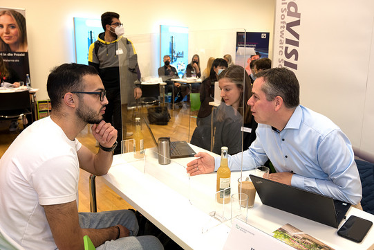 Ein internationaler Studierender sitzt an einem Tisch zwei Mitarbeitenden eines Unternehmens gegenüber. Einer der Unternehmensmitarbeiter erklärt etwas.